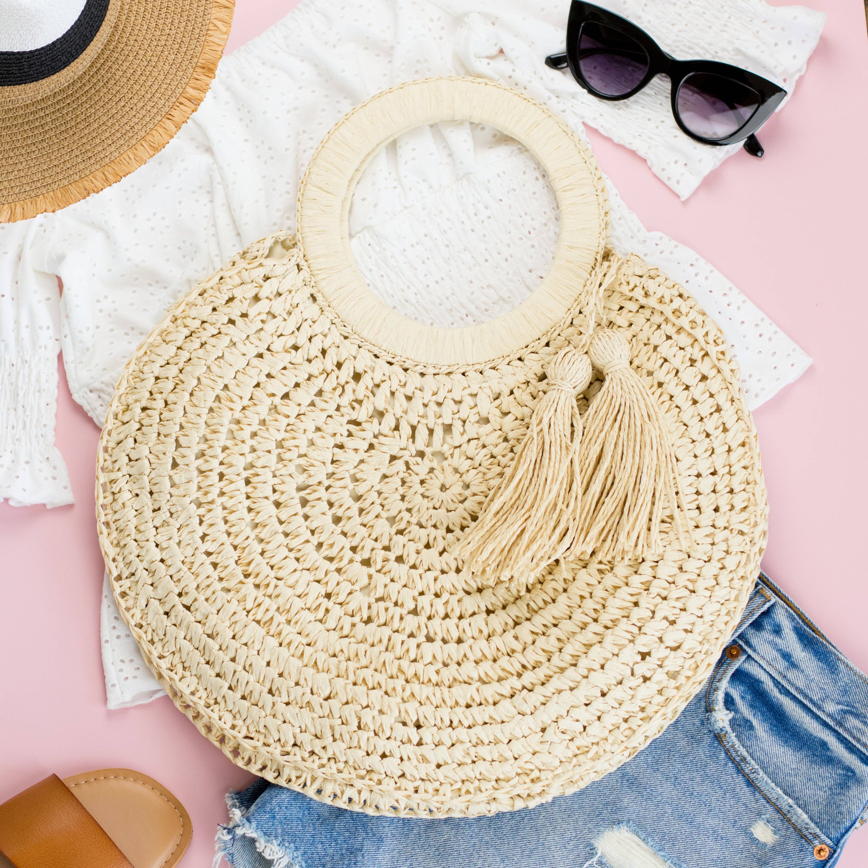 Woven straw bag with tassel on a pink surface with sunglasses and denim jeans.