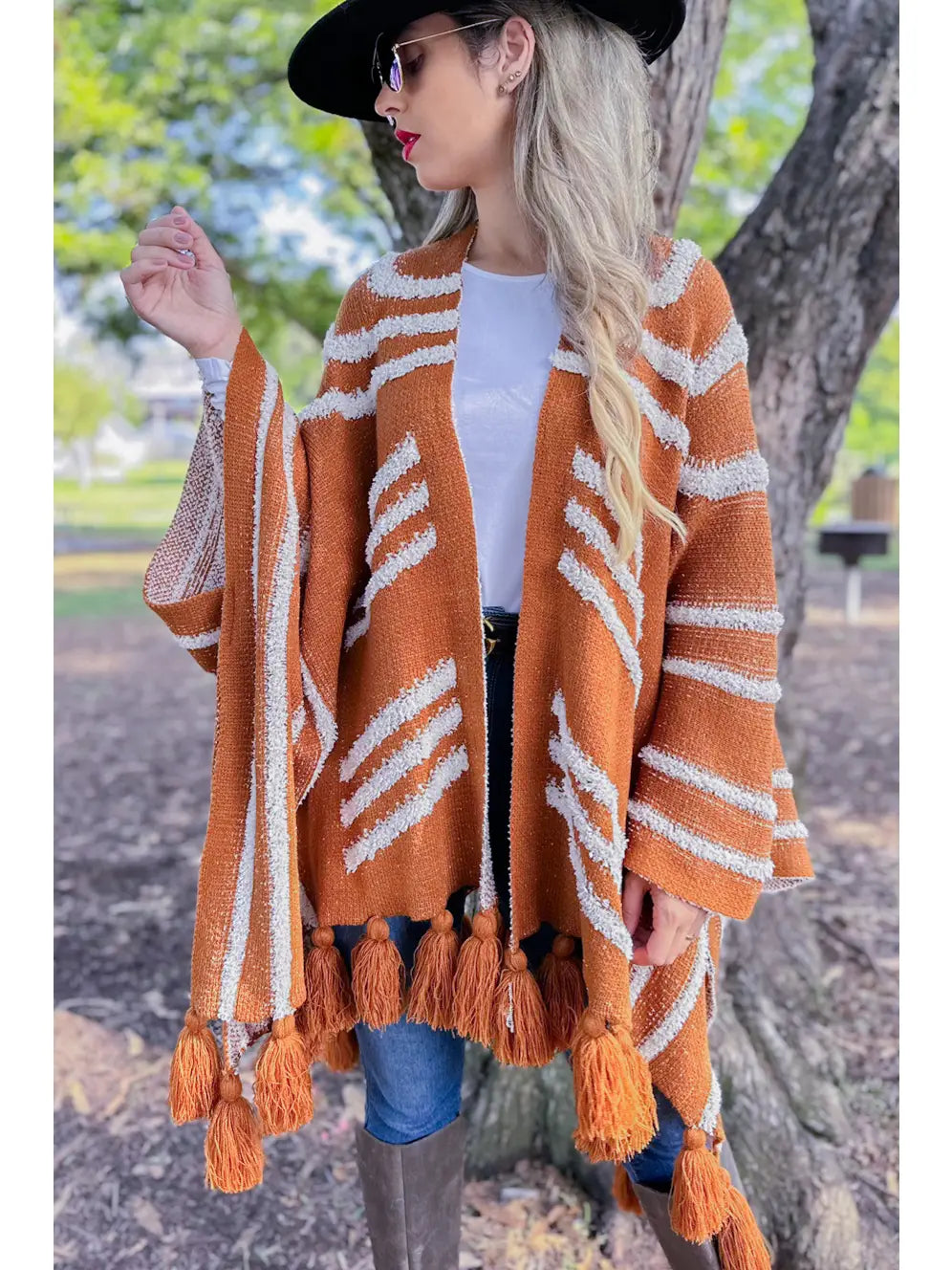 A woman wearing a rust-orange and white patterned knit poncho cardigan with tassels, styled with a white t-shirt, blue jeans, brown boots, and a black hat, standing outdoors in front of a tree.