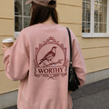 Person wearing a pink sweatshirt with a bird design and 'Worthy' text, holding a coffee cup.