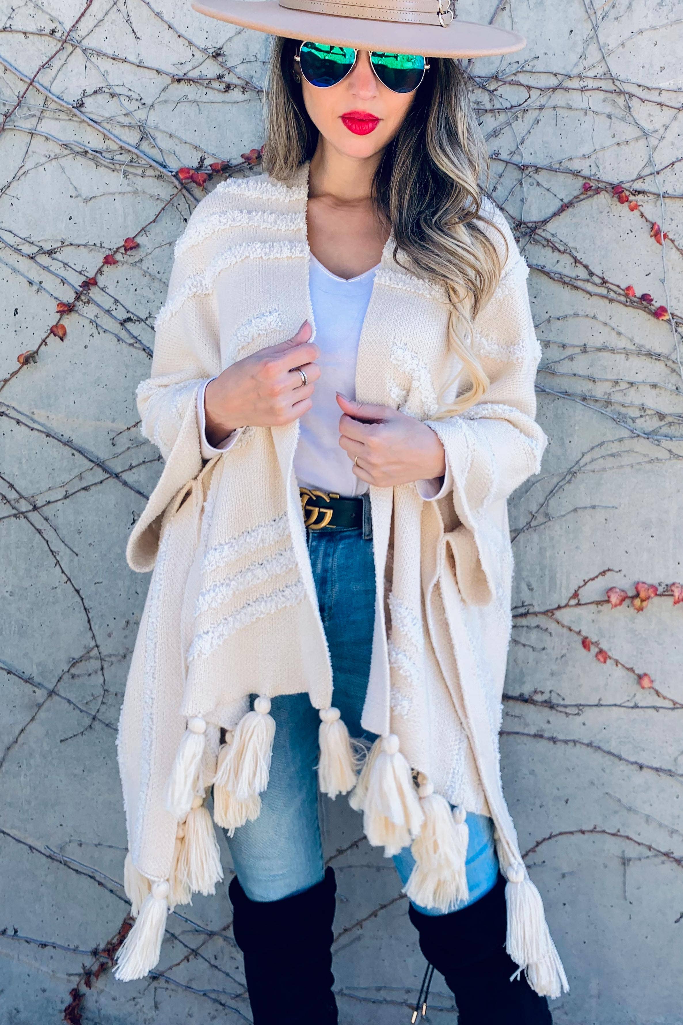 A woman wearing a cream knit poncho with white embroidered stripes and tassels, a white top, blue jeans, black over-the-knee boots, and a wide-brim beige hat, standing against a gray concrete wall with leafless vines.