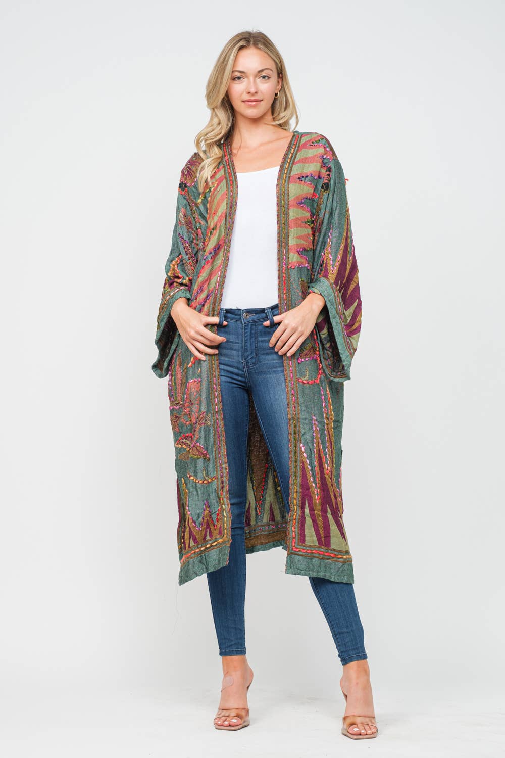 A woman modeling a long, open-front bohemian-style cardigan with a multicolor geometric and tribal-inspired print in green, red, orange, and blue, worn over a white top and blue jeans against a light gray studio background.