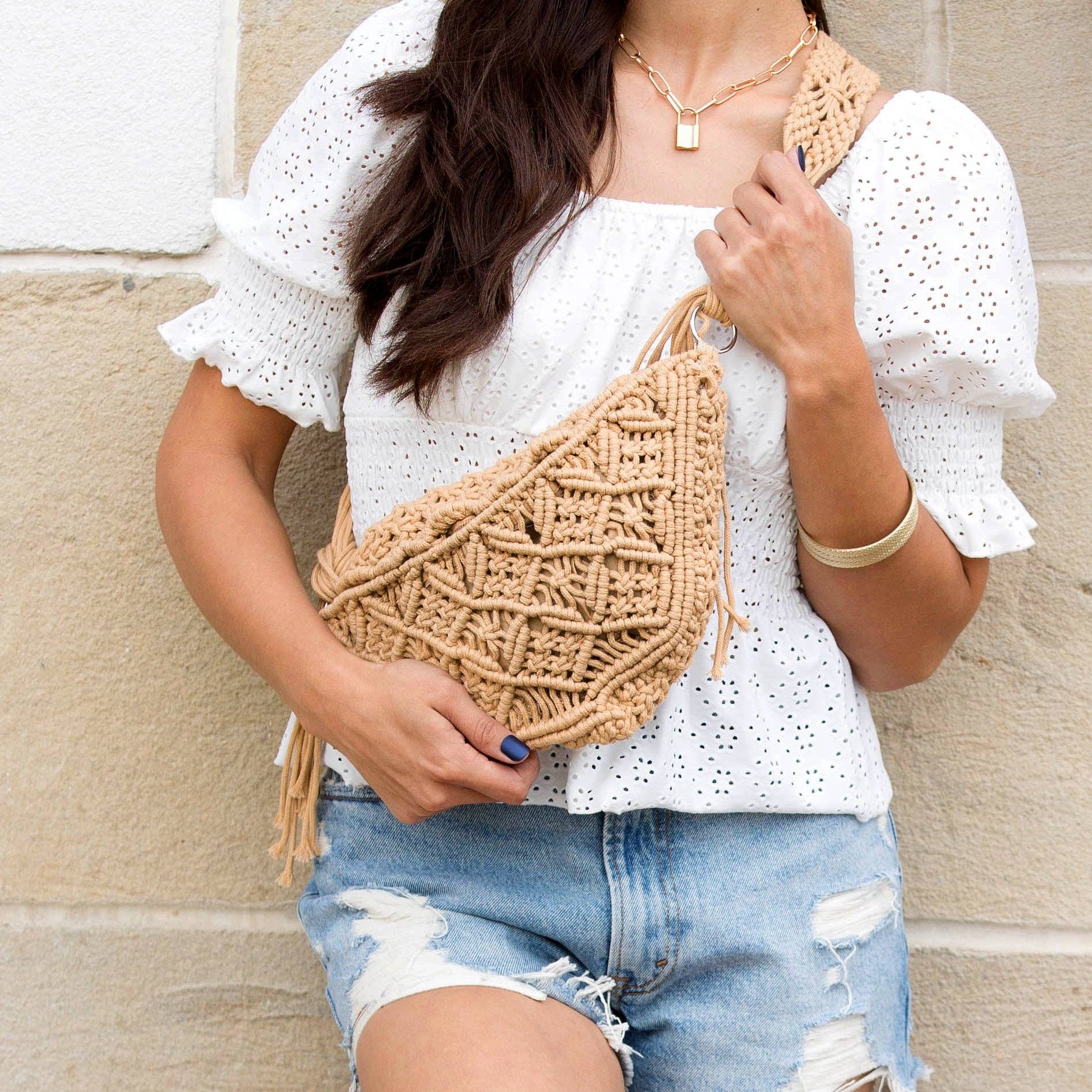 Person wearing a white blouse and denim shorts, holding a beige woven bag against a beige wall.