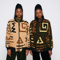 Two models wearing patterned knit bomber jackets against a plain light gray studio background. The jacket on the left is beige with black and brown geometric motifs, and the jacket on the right is dark olive green with cream, brown, and black geometric motifs.