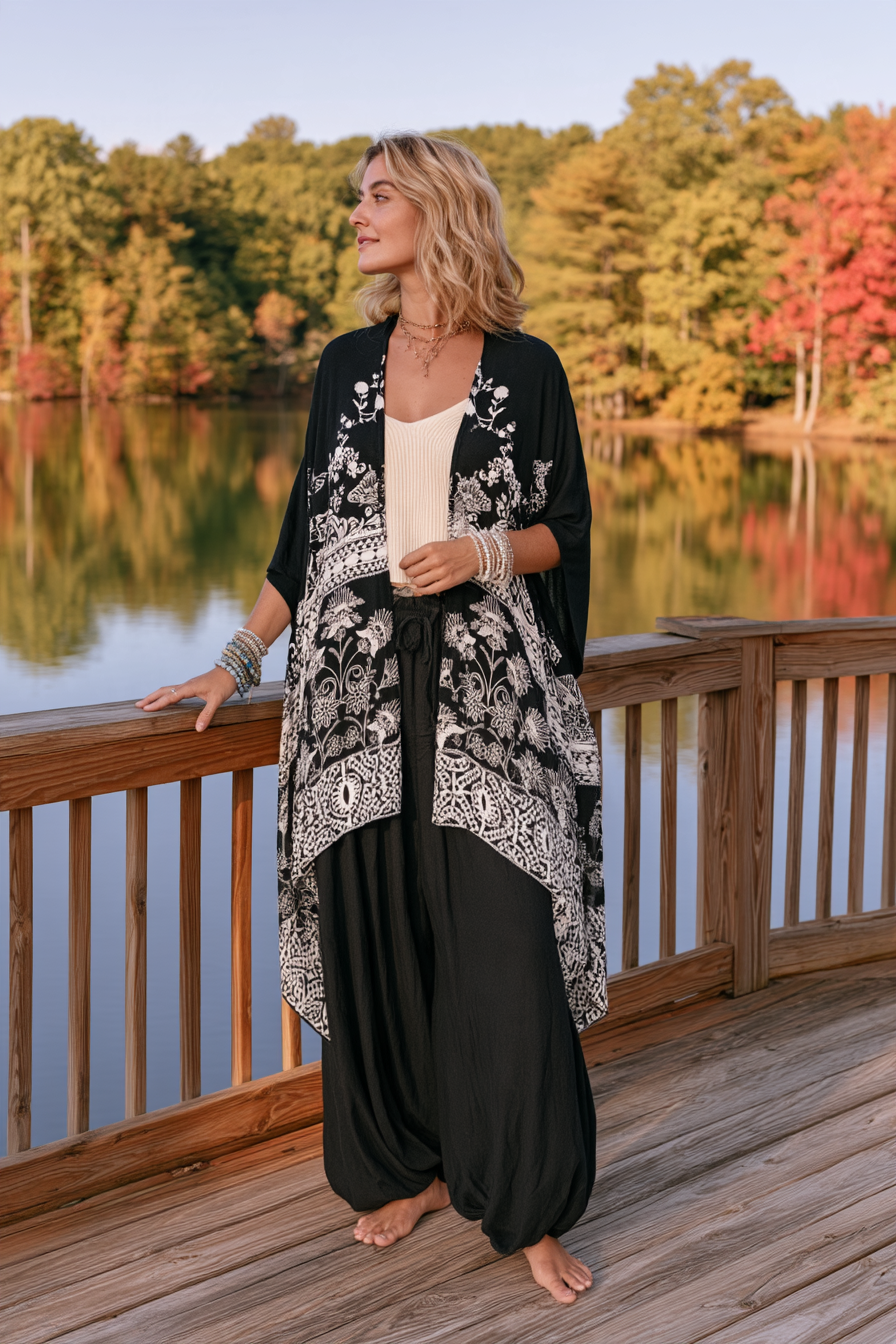 A woman stands barefoot on a wooden deck beside a lake, wearing a black and white patterned open-front cardigan over a cream top and black harem pants. Trees with autumn colors are reflected in the water behind her.