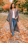 A woman stands on a leaf-covered path in an autumn park wearing a long, open-front patterned kimono cardigan over a white top and blue jeans.