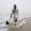 A woman walks a small dog on a leash along a sandy beach with ocean waves in the background, wearing a hooded one-piece jumpsuit with a large floral print in beige, blue, and pink.