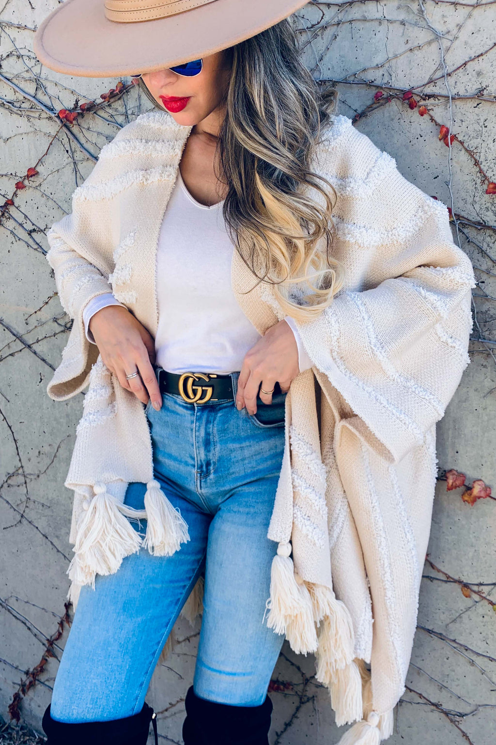 A woman wearing a cream knit poncho with fringe, a white top, light blue jeans, a black Gucci belt, and a wide-brim tan hat, posing against a textured stone wall with climbing vines.